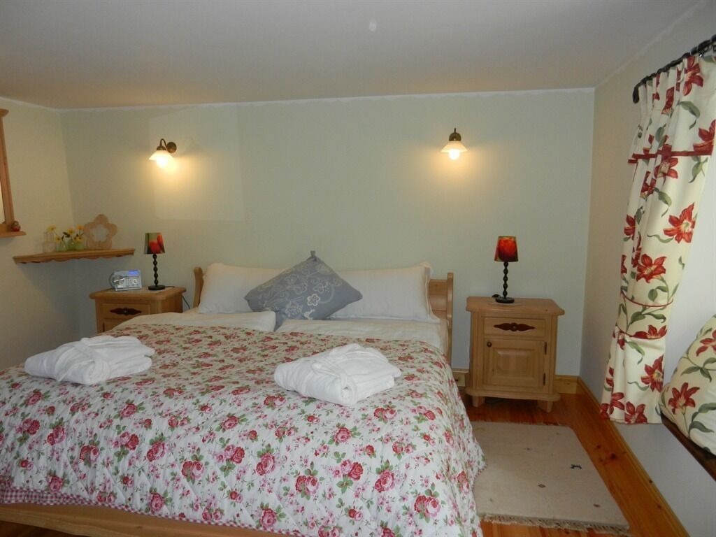 A bedroom with a floral bedspread, two bedside lamps, a nightstand with a radio, and a window with floral curtains.