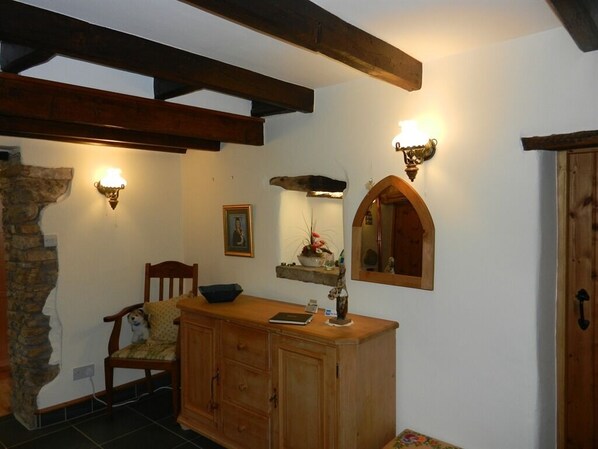 Una habitación rústica con vigas de madera a la vista, una pared de piedra, un armario de madera, una silla, una mesita y luces fijadas en la pared.