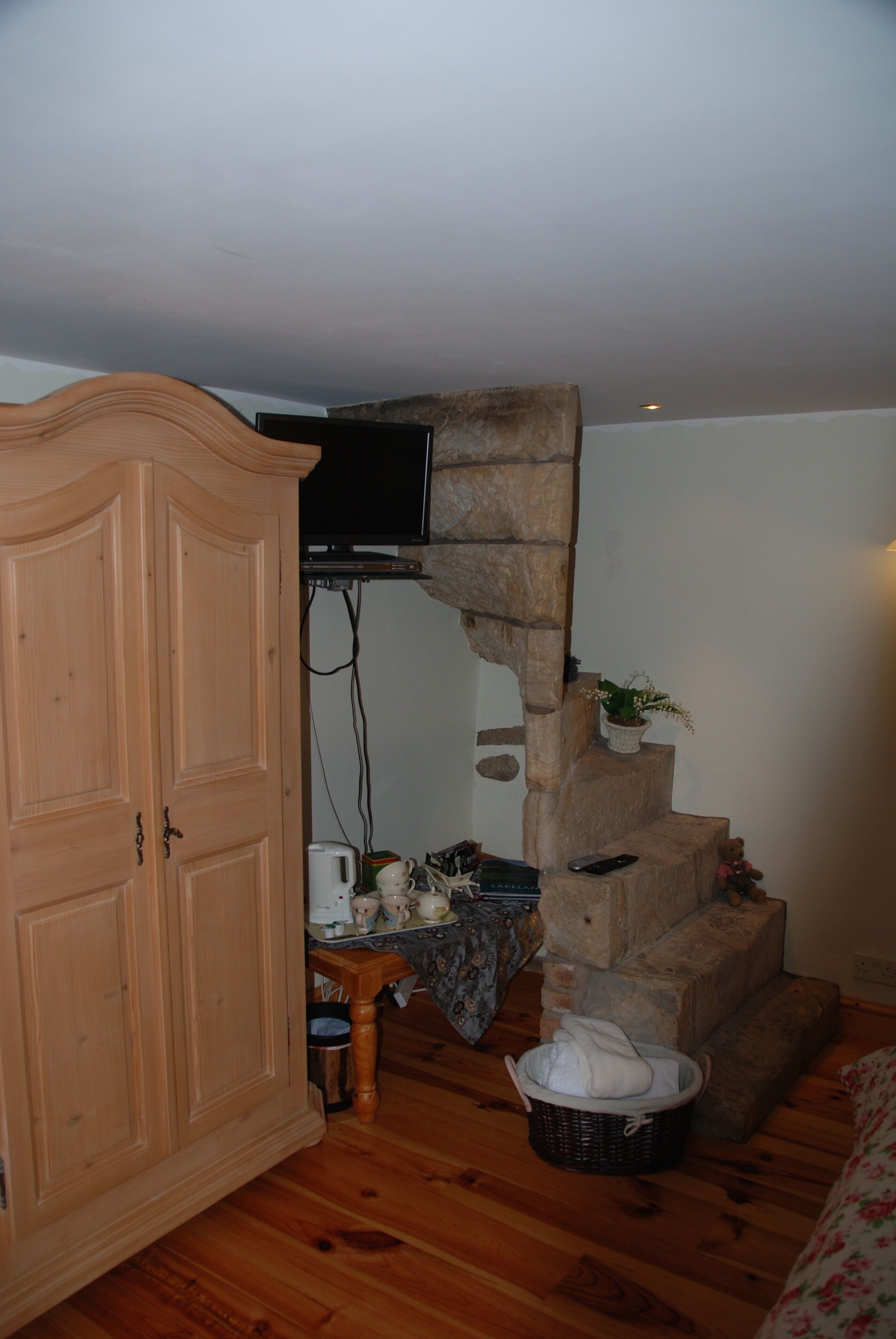 A room with a wooden wardrobe, a stone staircase, a television mounted on the wall, a small table with a basket, and a teddy bear.