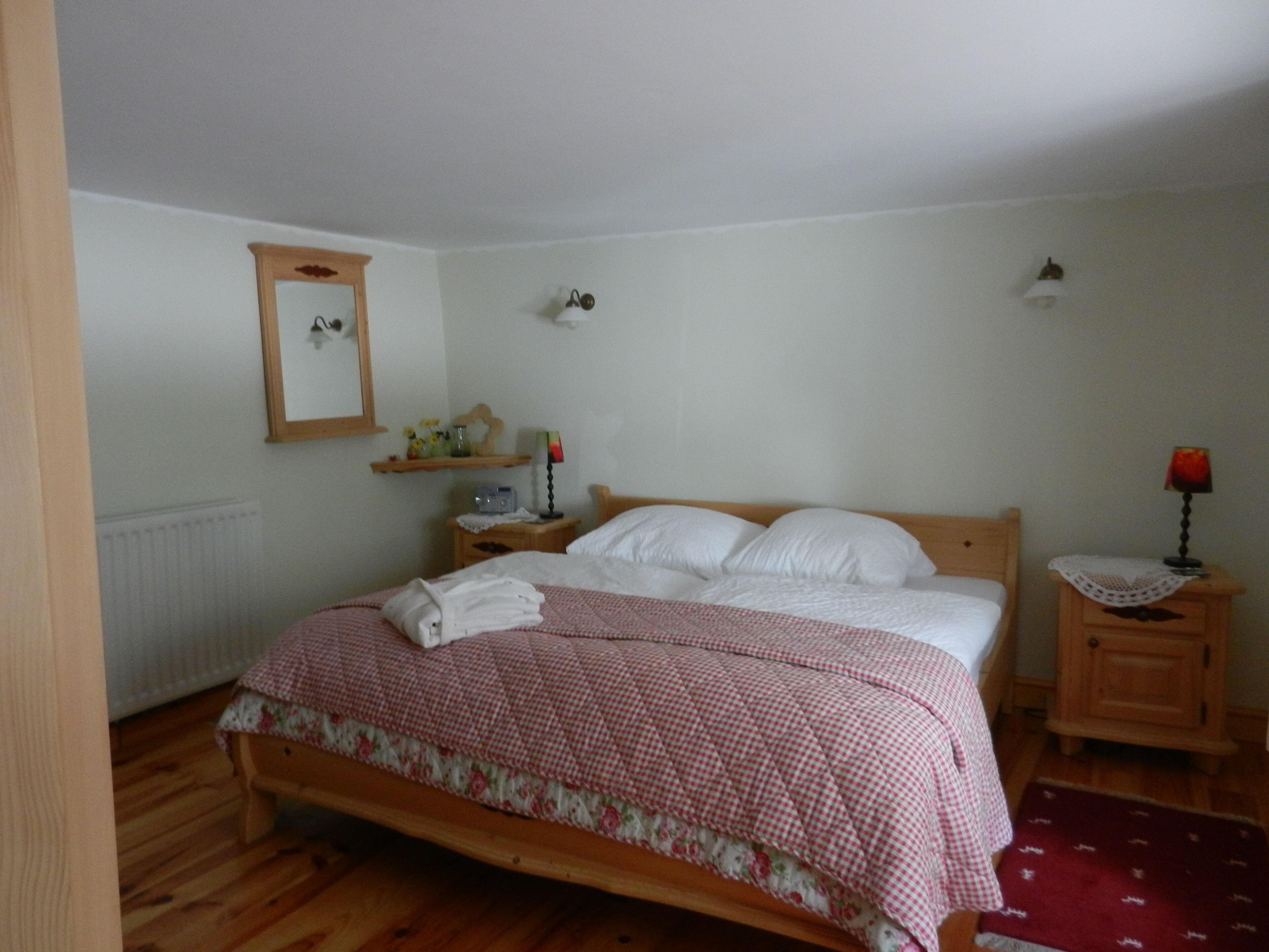 Un dormitorio con cama de madera, almohadas blancas, colcha a cuadros rojo y blanco, mesita de noche de madera con una lámpara, espejo en la pared y un radiador.