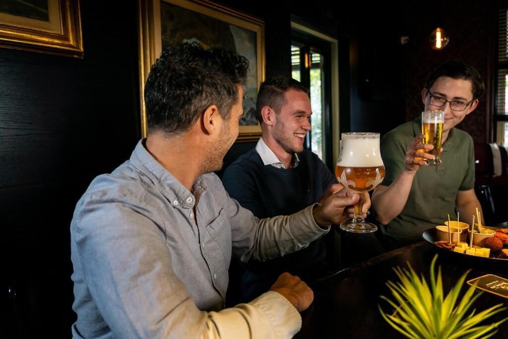 Drei Personen sitzen an einer Bar, vor ihnen Biergläser, eine Platte mit Snacks und ein Topf mit einer Pflanze auf dem Tisch.