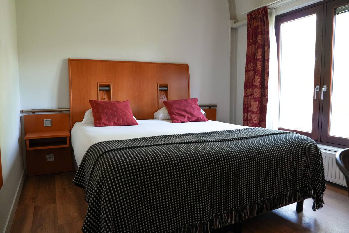 A hotel room with a wooden headboard, two red pillows, a black and white patterned bedspread, a wooden nightstand, and a window with a red curtain.