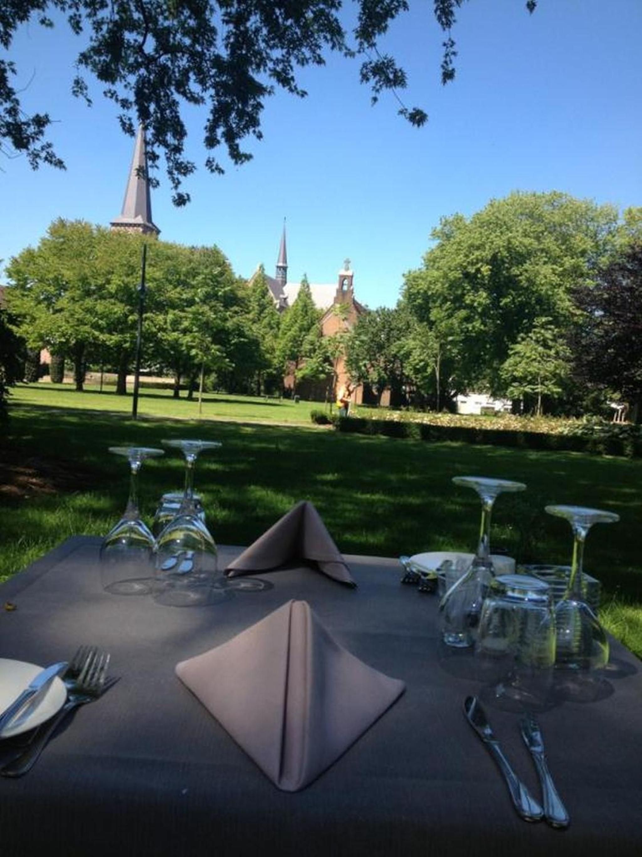 An outdoor dining table set with glasses, cutlery, and napkins, with a church and trees in the background.