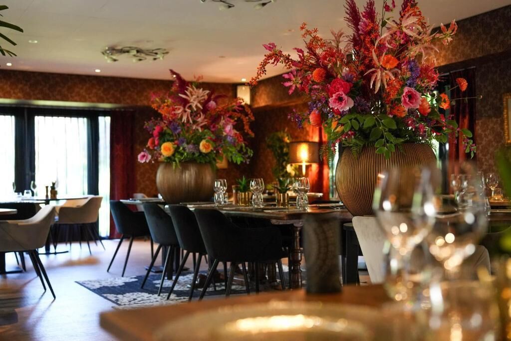 Un coin repas avec une table dressée pour deux personnes, entourée de chaises, et de grandes compositions florales en arrière-plan.