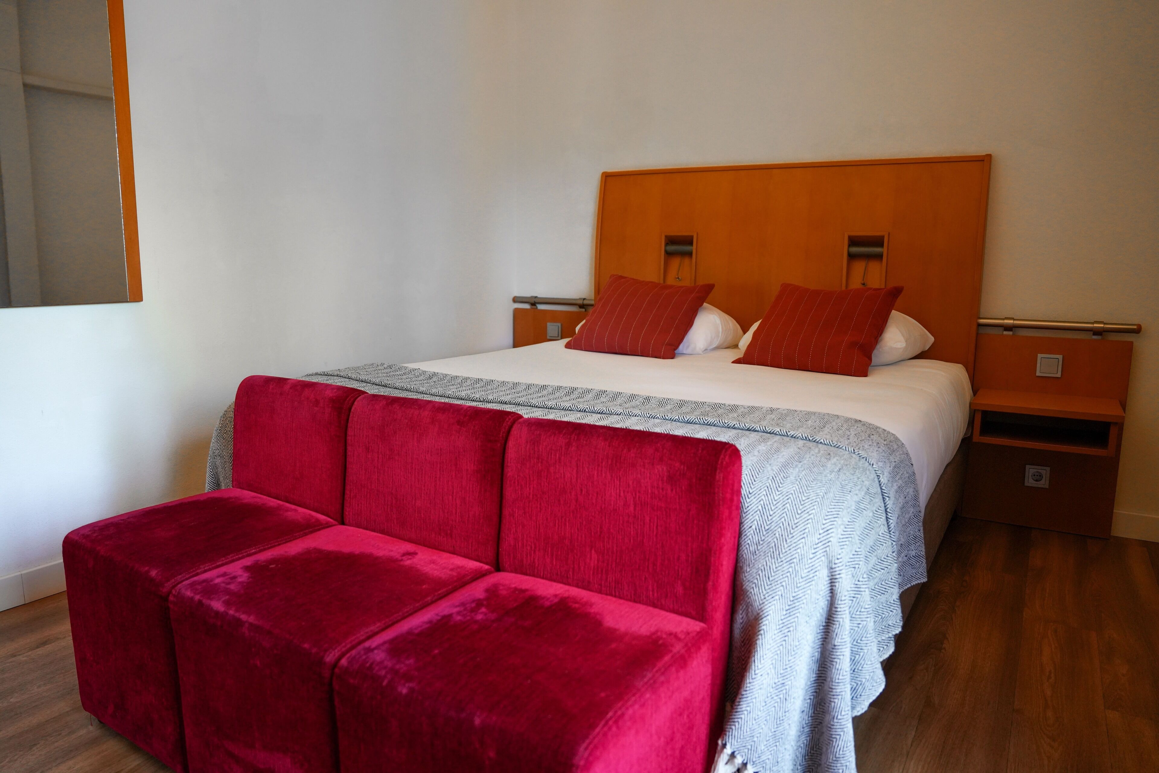 A neatly made bed with a wooden headboard, two bedside tables, a red upholstered bench, and a patterned throw blanket.