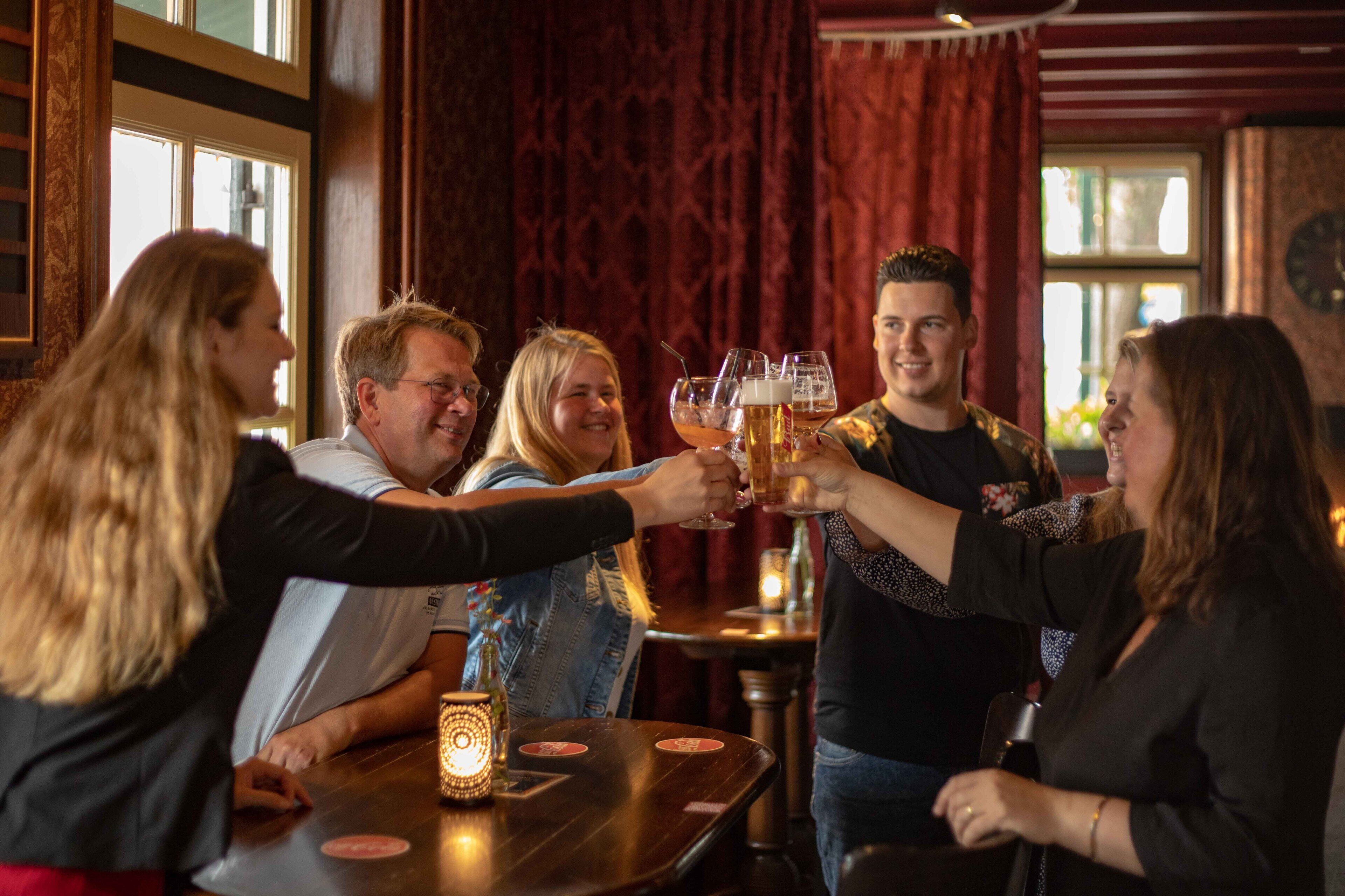 Een groep mensen die met bierglazen proosten in een bar met houten meubels en een kaars op tafel.