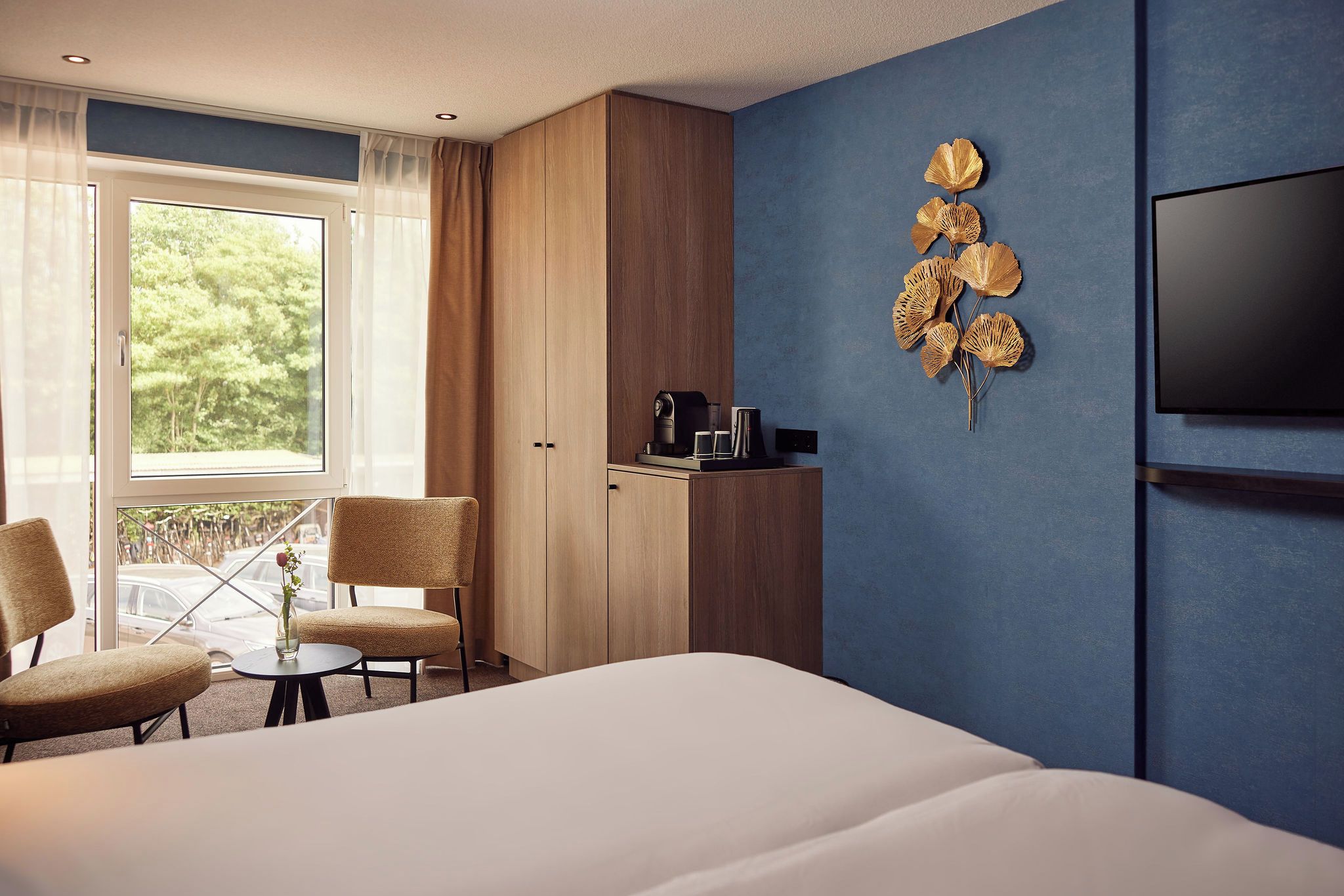 A hotel room with a bed, a desk with a coffee maker, chairs, a window with curtains, and a wall-mounted TV.