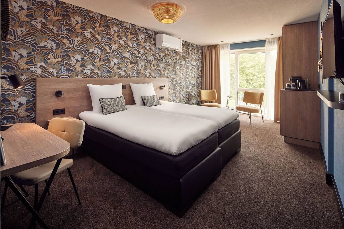 A modern hotel room with a large bed, a desk with a chair, a small table, and a chair. There is a window with curtains and a view of greenery outside.