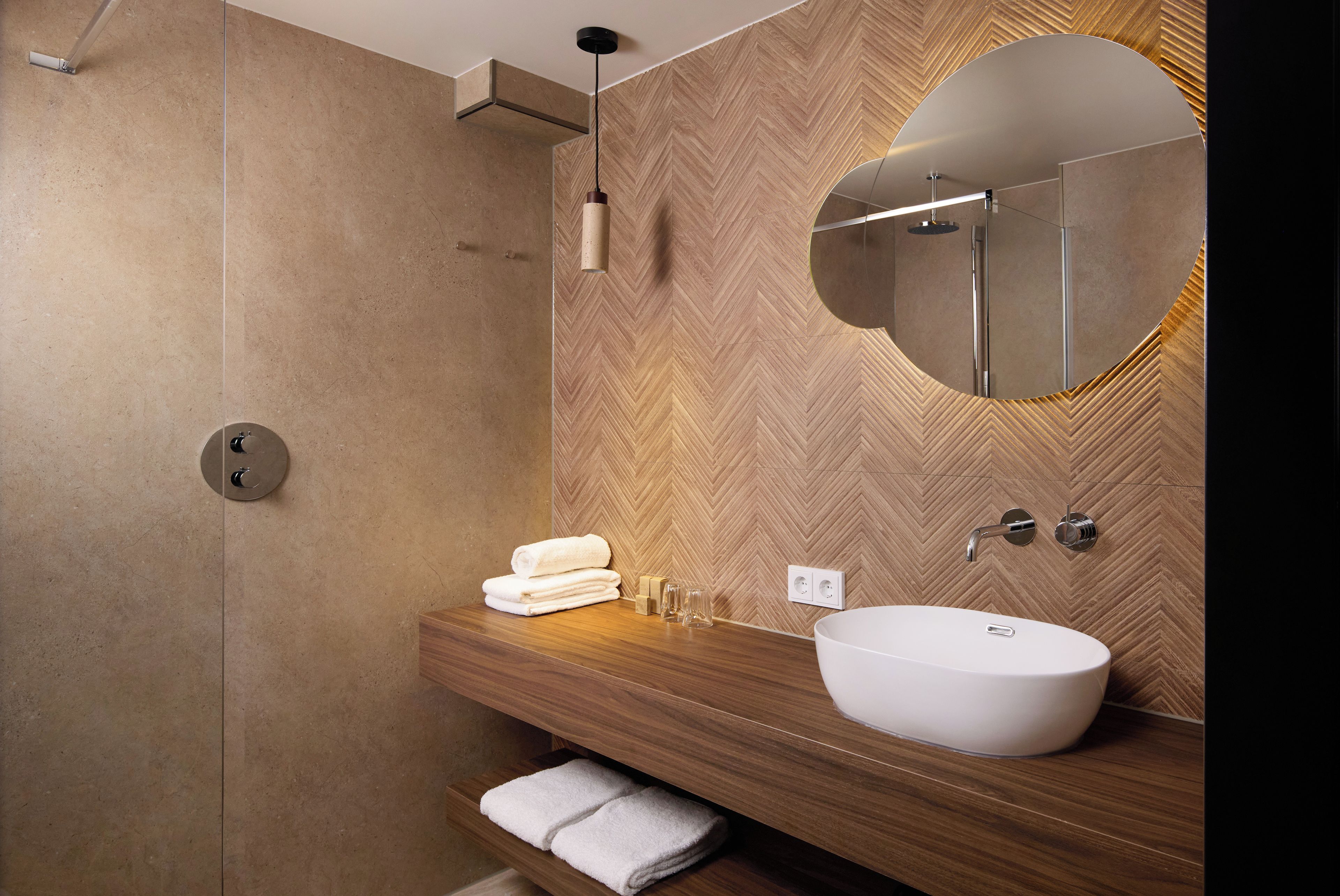 A modern bathroom with a wooden herringbone wall, a round mirror, a white sink, and a glass shower enclosure.