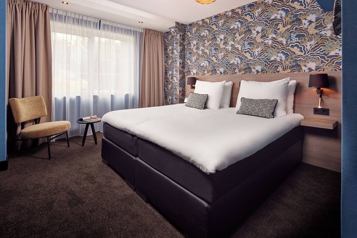 A hotel room with a large bed, a chair, a small table, and a patterned wall.