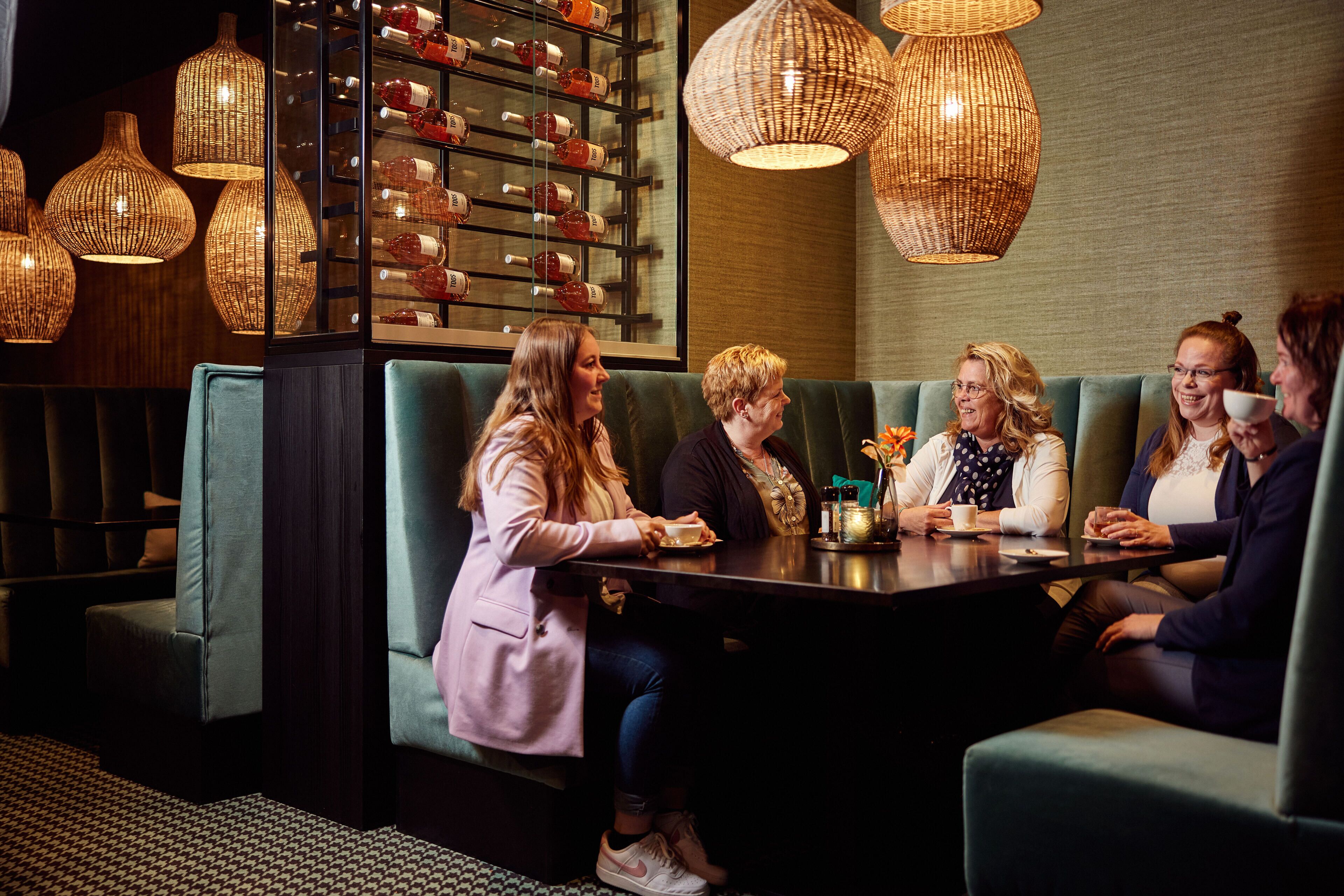 Un grupo de personas sentadas alrededor de una mesa en una habitación acogedora, con luz tenue, lámparas colgantes de mimbre y una estantería para vinos al fondo.