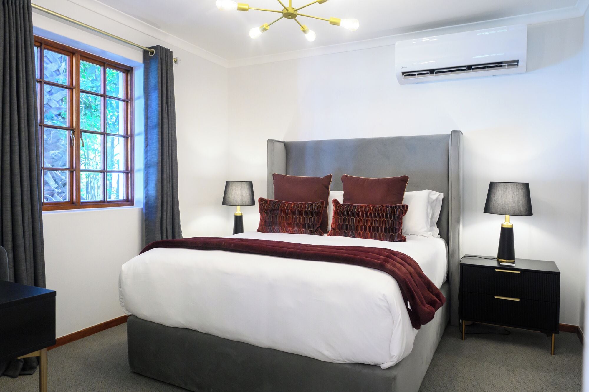A hotel room with a large bed, two bedside tables with lamps, a ceiling light fixture, and an air conditioning unit.