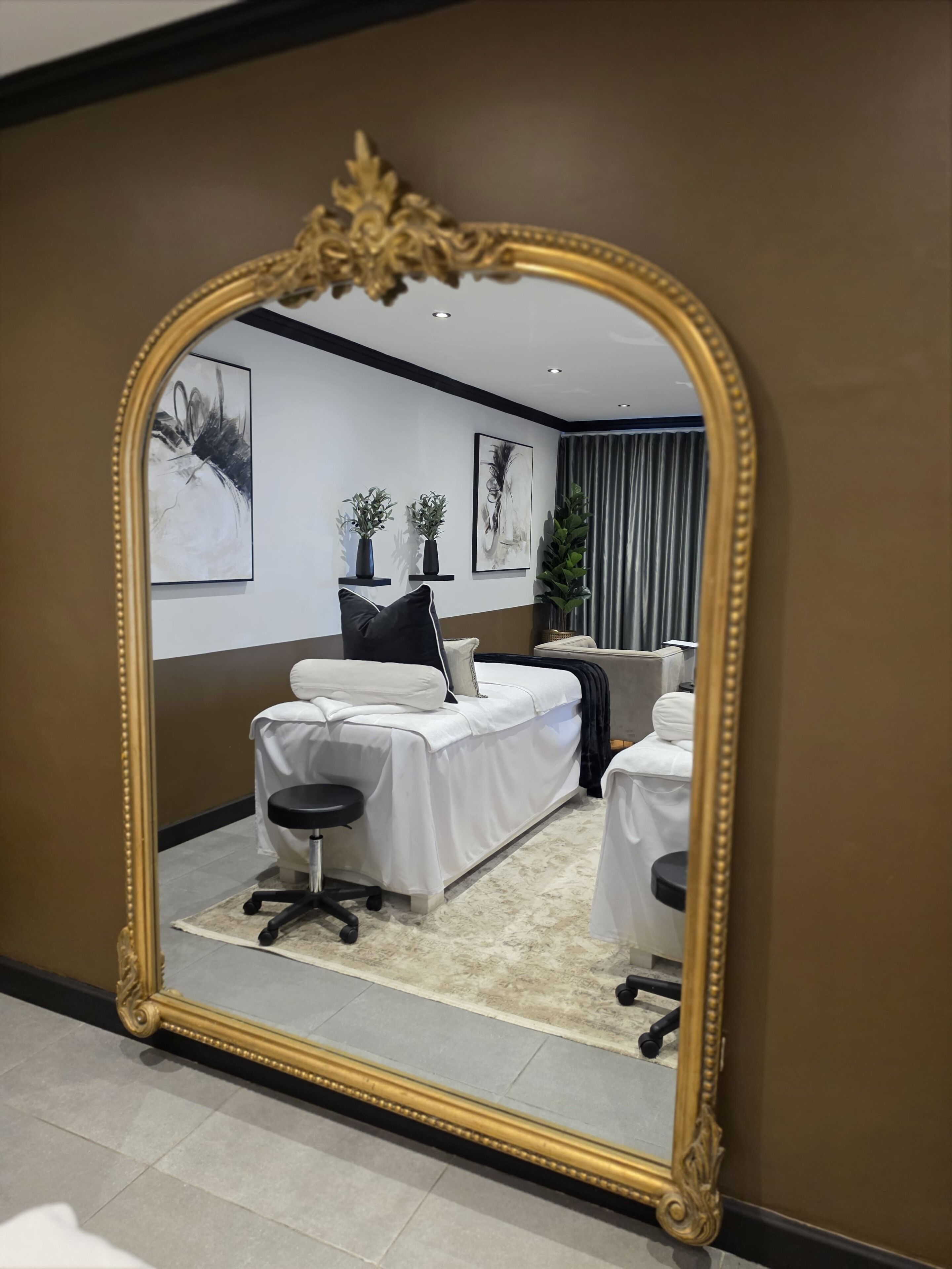 A room with a large ornate mirror, a bed, a chair, and two small tables.
