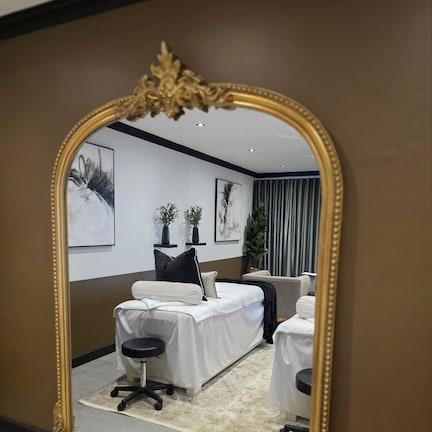 A room with a large ornate mirror, a bed, a chair, and two small tables.