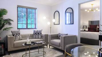 A living room with a sofa, armchairs, a coffee table, and a glass table. There are two wall-mounted mirrors and a ceiling light fixture.