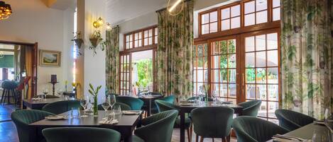 Un ristorante con tavoli in legno e sedie verdi, ampie finestre con tende e piante pensili.