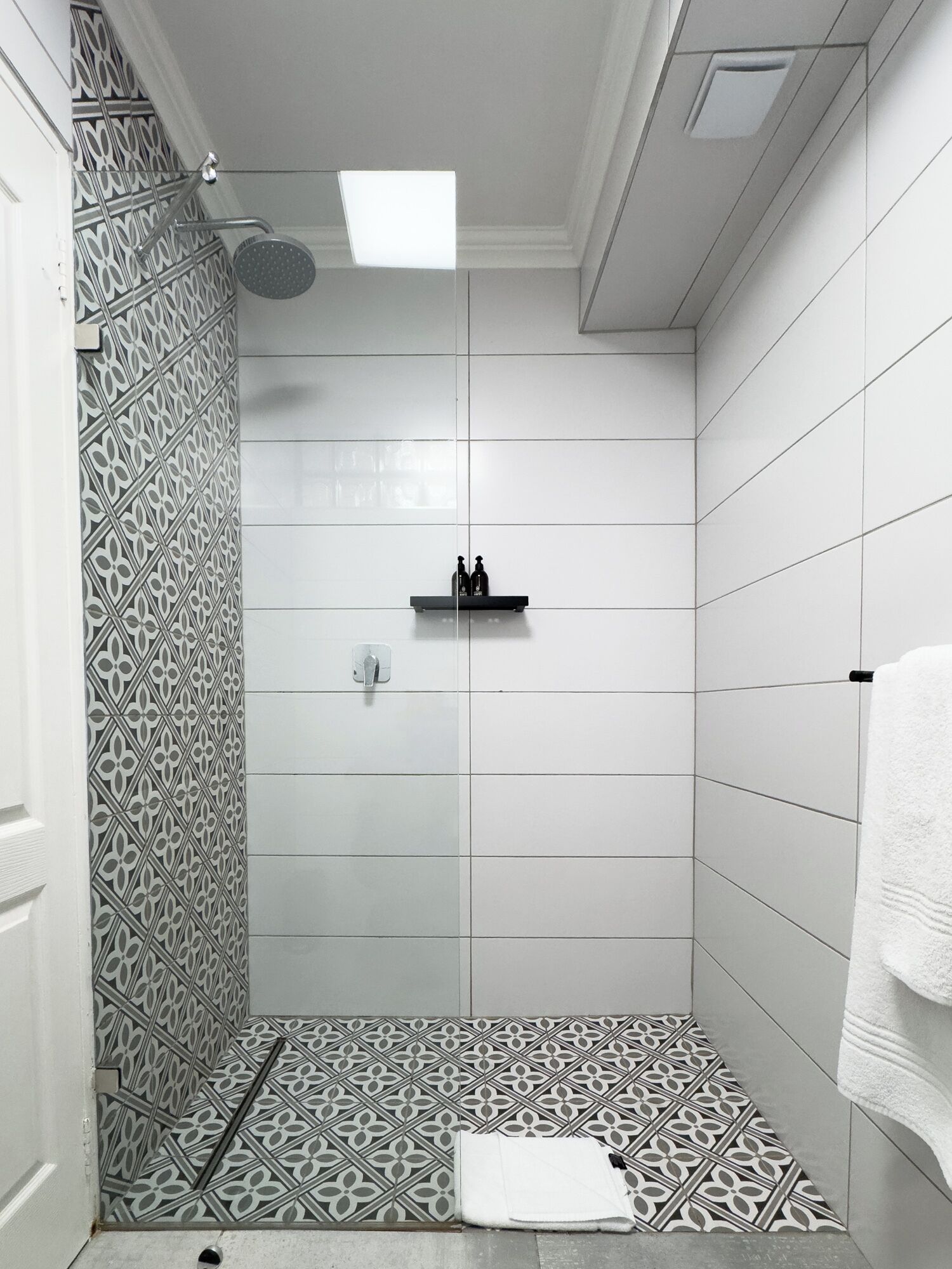 A shower with a patterned tile wall, a shelf with bottles, and a white towel on the floor.