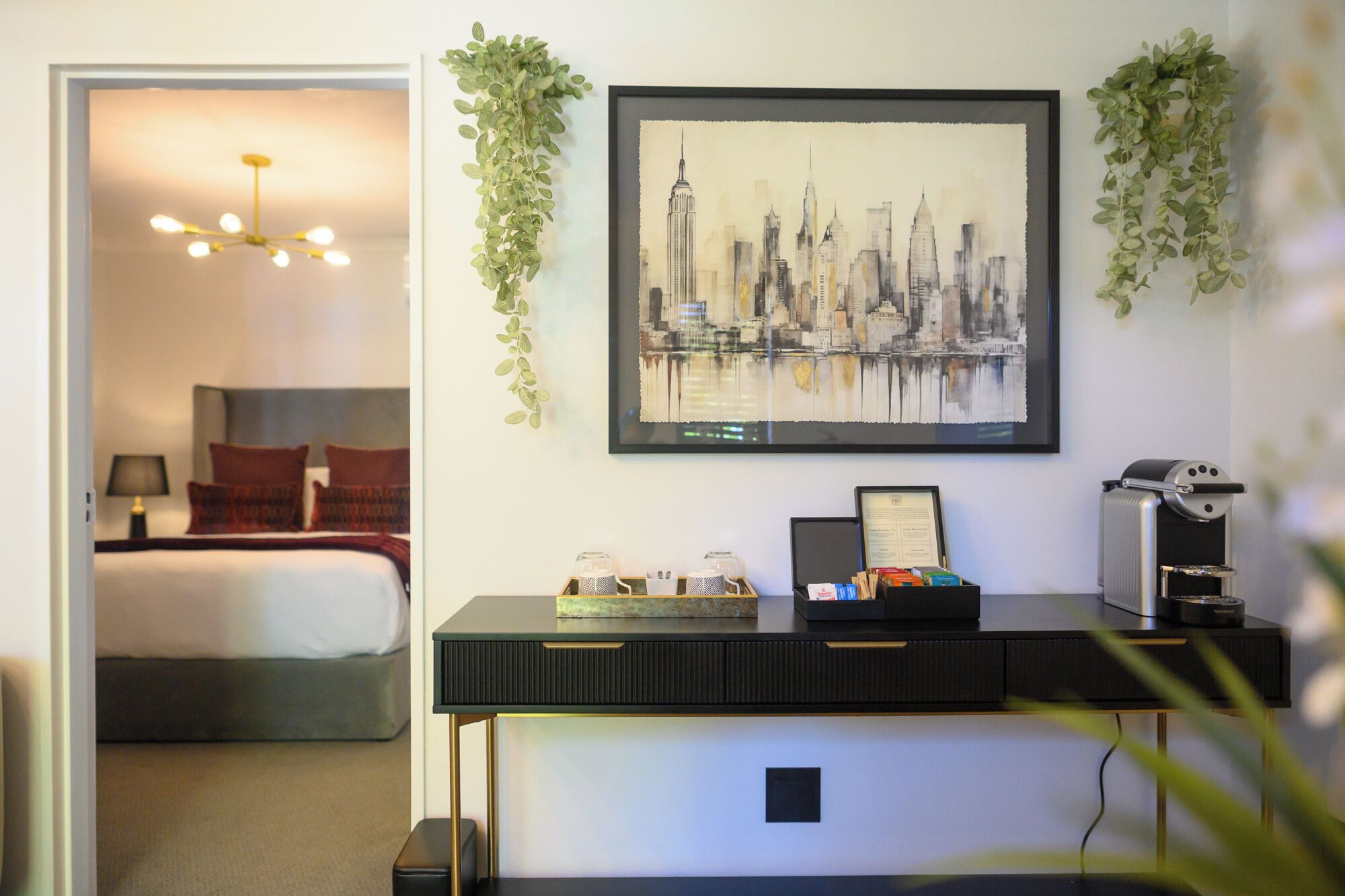 A hotel room with a bed, a desk with a coffee machine, a framed cityscape painting, and hanging greenery.