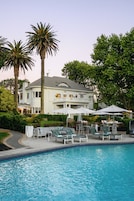 A pool area with lounge chairs and umbrellas, a large house with a balcony, and palm trees.
