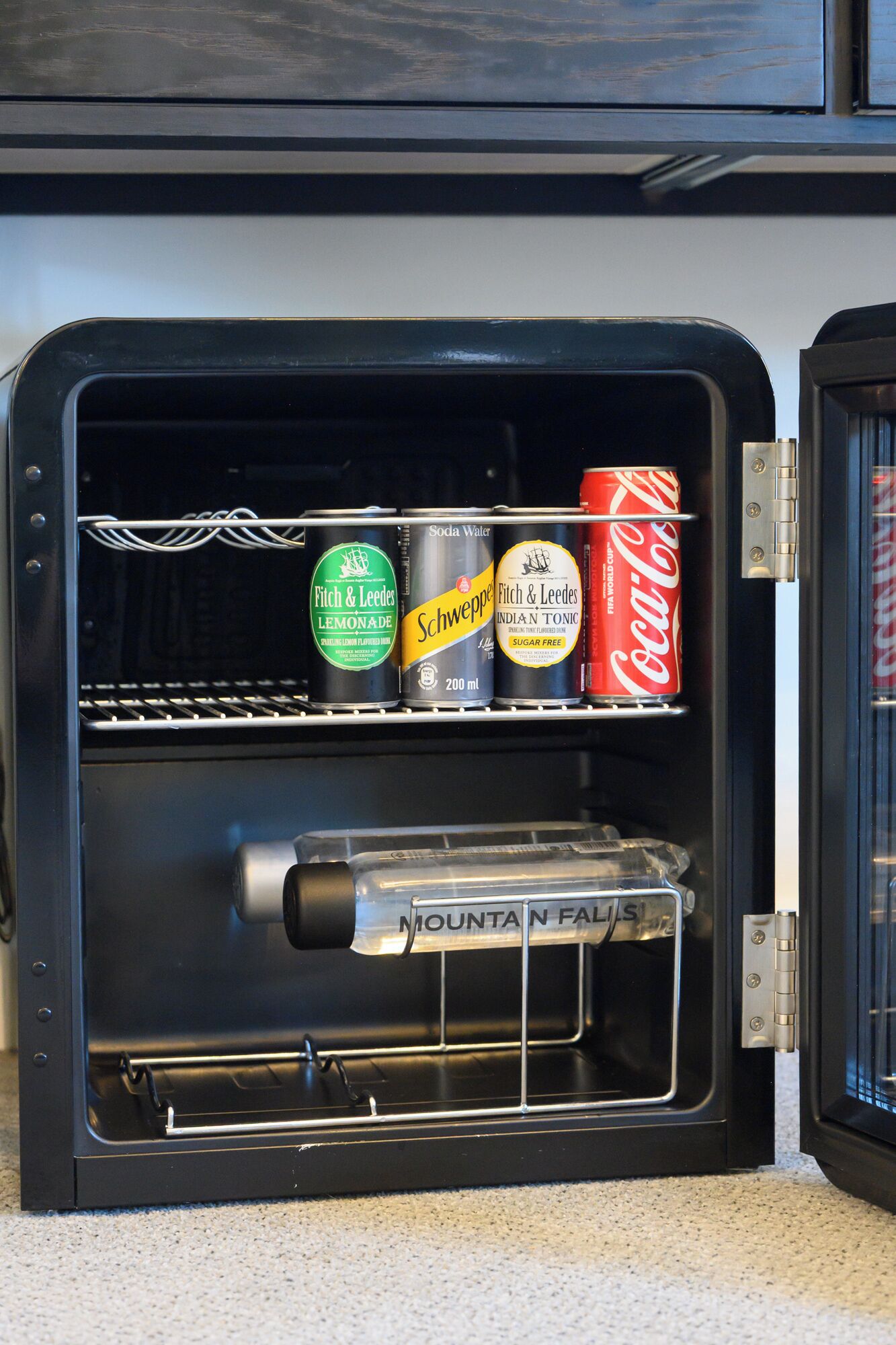 Eine Minibar mit Getränken, darunter Sprudelwasser, Zitronenlimonade, Tonic Water und eine Dose Coca-Cola.