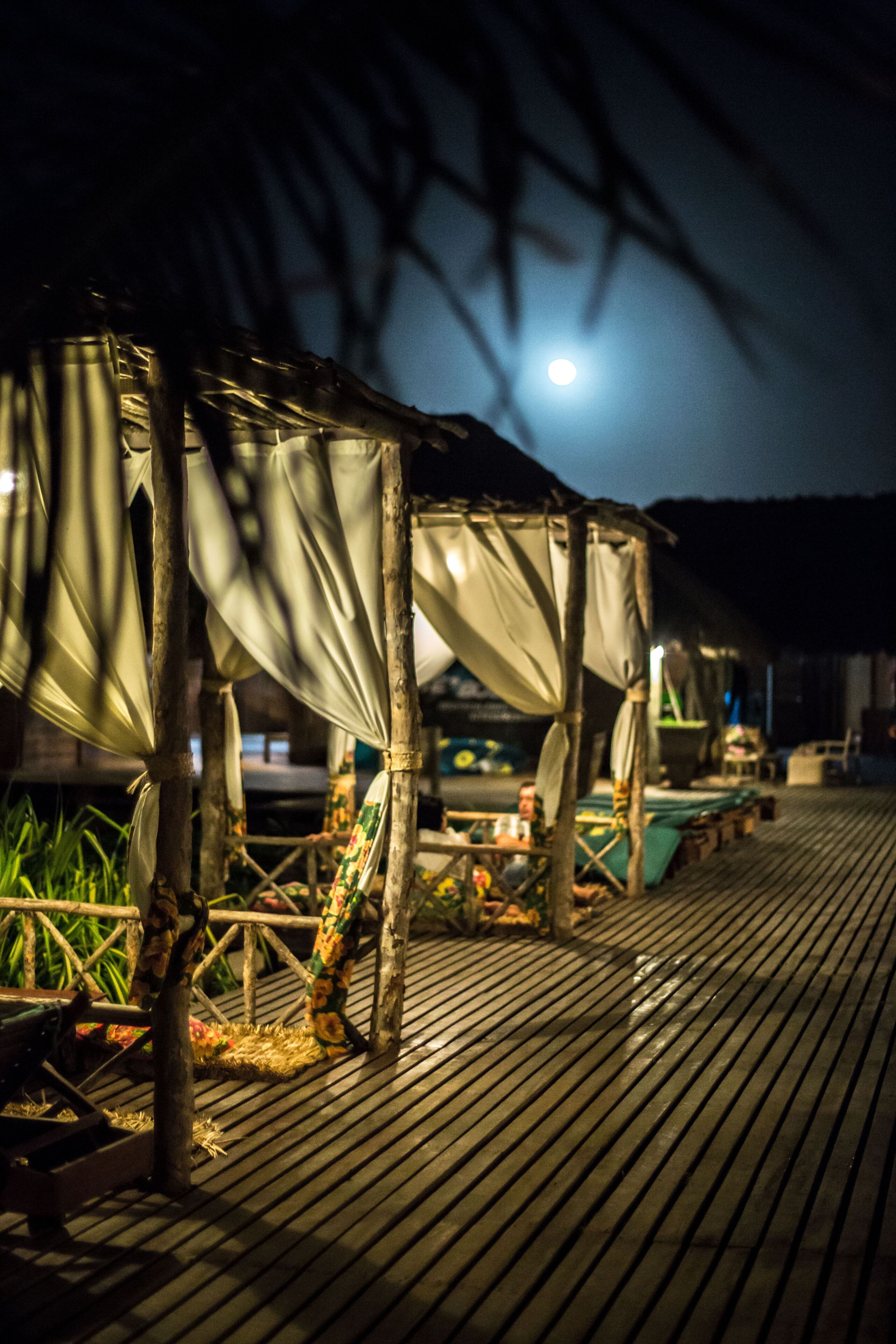 Une terrasse en bois surmontée d’une structure formant auvent, probablement aménagée pour les repas ou la détente en plein air, sous un ciel nocturne où l’on peut apercevoir la lune.