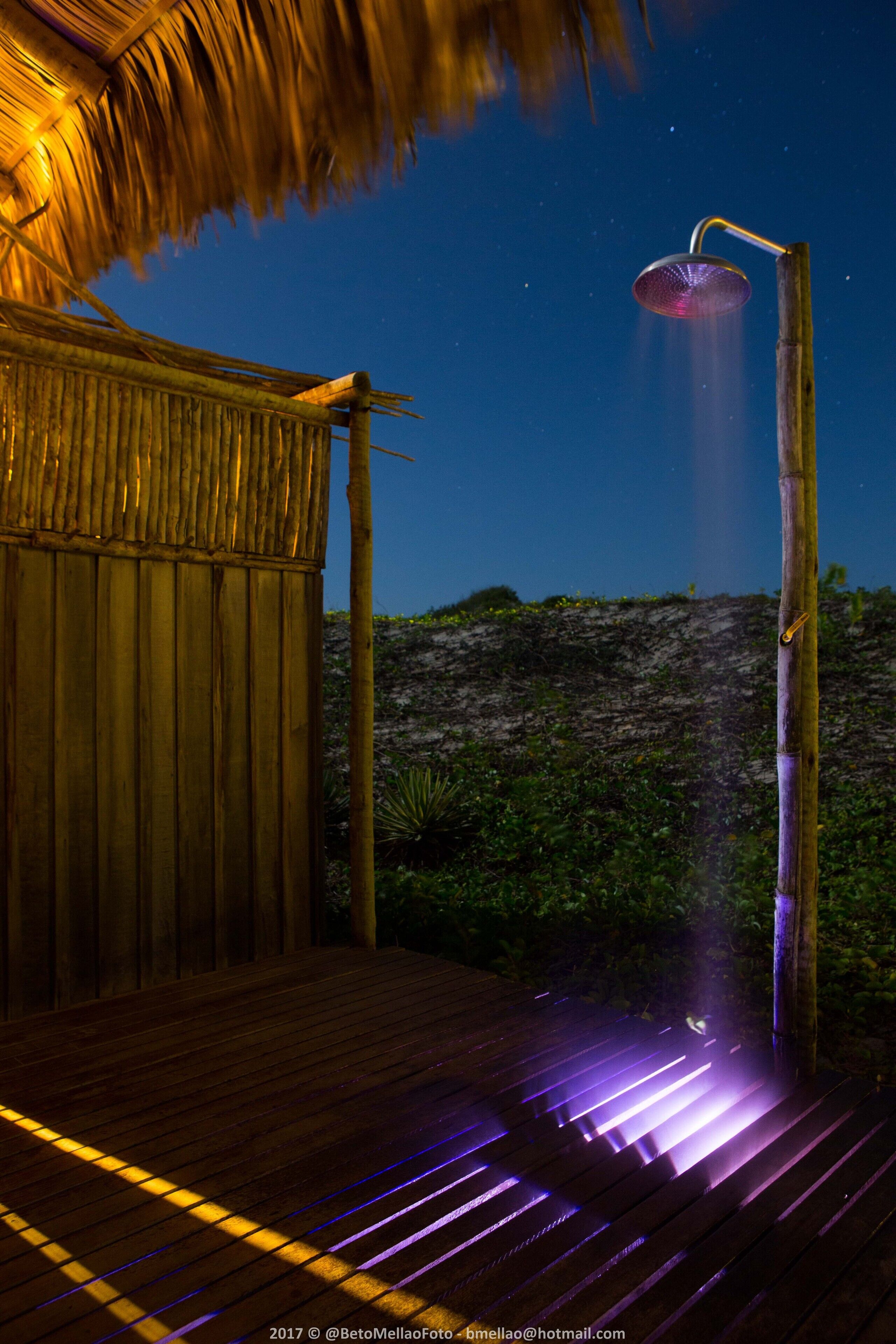 A wooden deck with a thatched roof structure, a bamboo shower, and a view of a rocky landscape.
