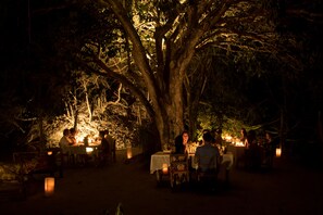 An outdoor dining area with tables set under a large tree, surrounded by lanterns.
