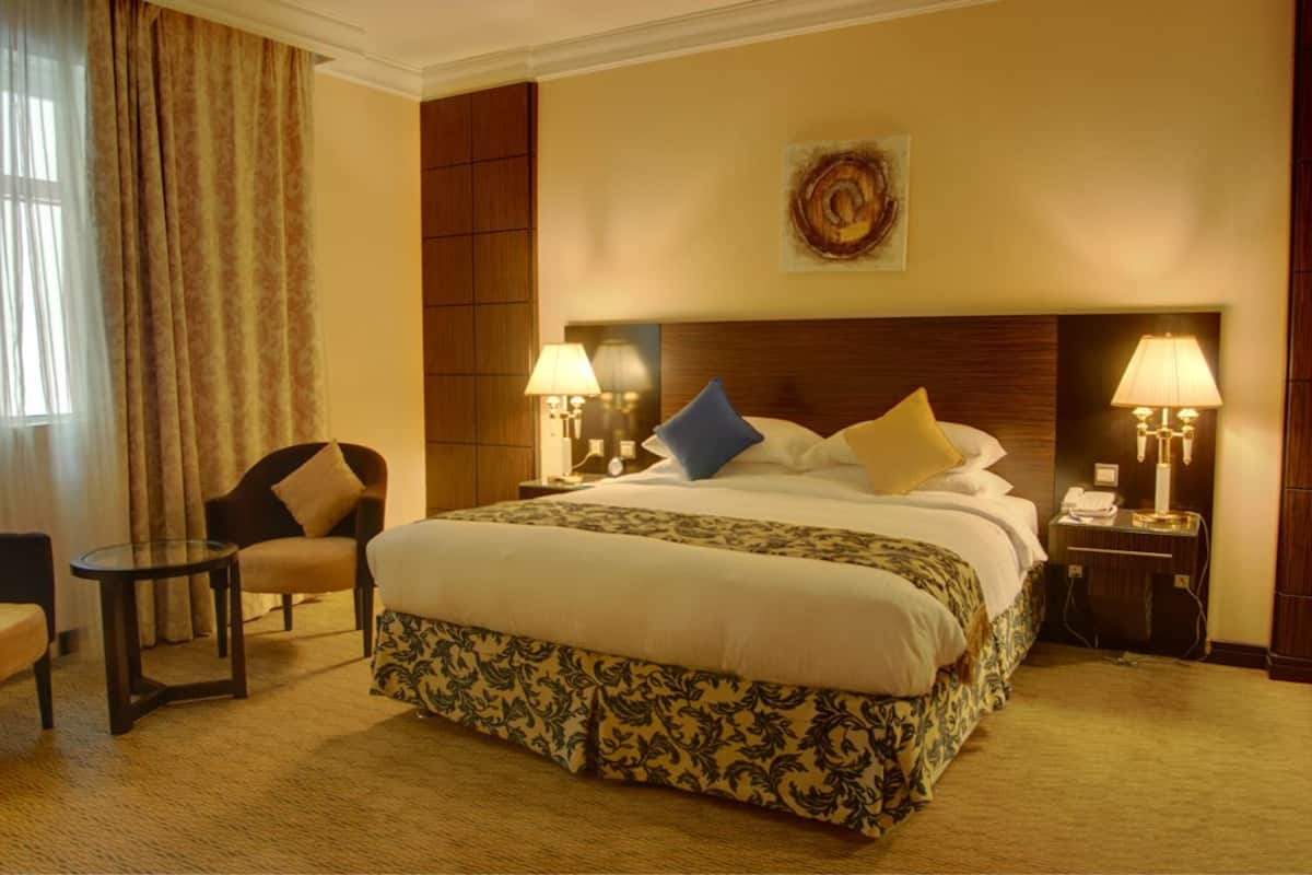 Een hotelkamer met een groot bed, twee nachtkastjes met lampen, een stoel, een klein tafeltje en een groot raam met gordijnen.