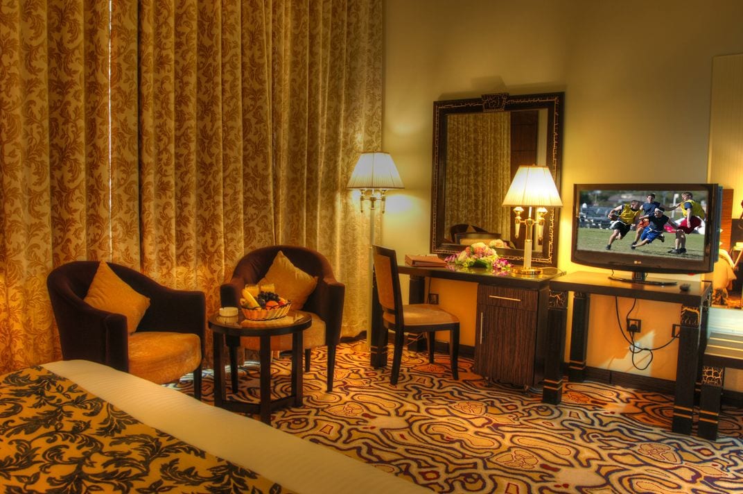 A hotel room with a large bed, two armchairs, a desk with a television, and a patterned carpet.
