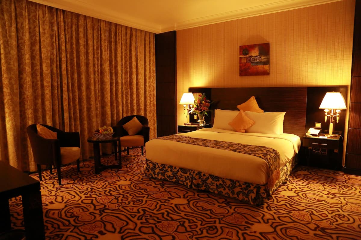 Een hotelkamer met een groot bed, twee nachtkastjes met lampen, een stoel en een kamerbreed tapijt met een patroon.