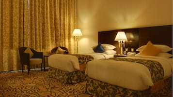 Quarto de hotel com duas camas, uma cadeira, uma mesinha e um abajur.