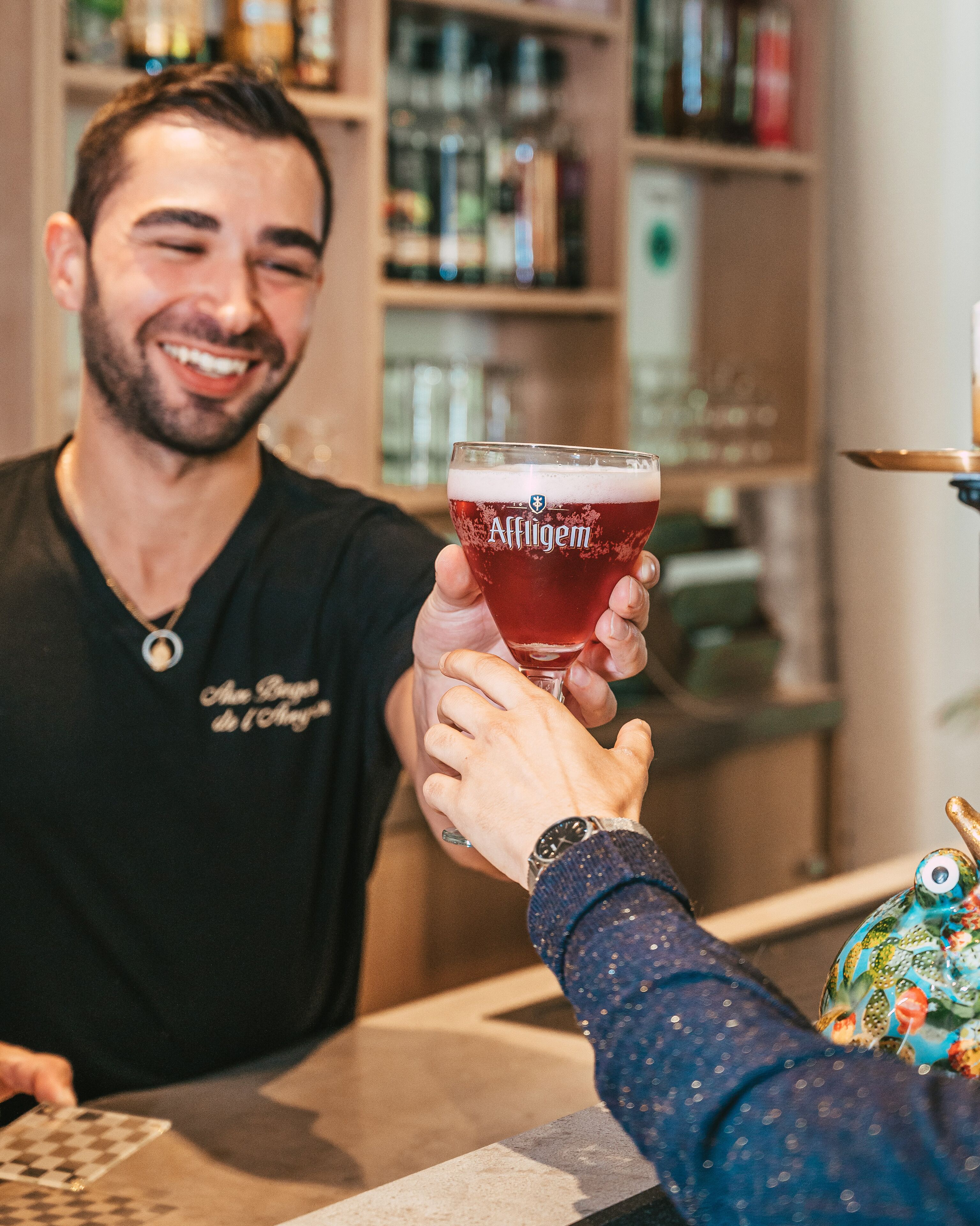 A person holding a glass of Affligem beer at a bar.