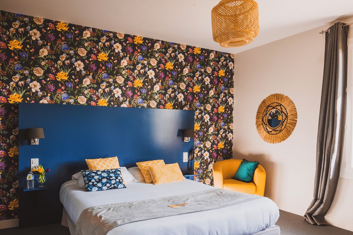 Une chambre avec un mur orné d’un motif floral, une tête de lit bleue, un lit recouvert d’une couverture grise, un fauteuil jaune et un luminaire en matière tressée.