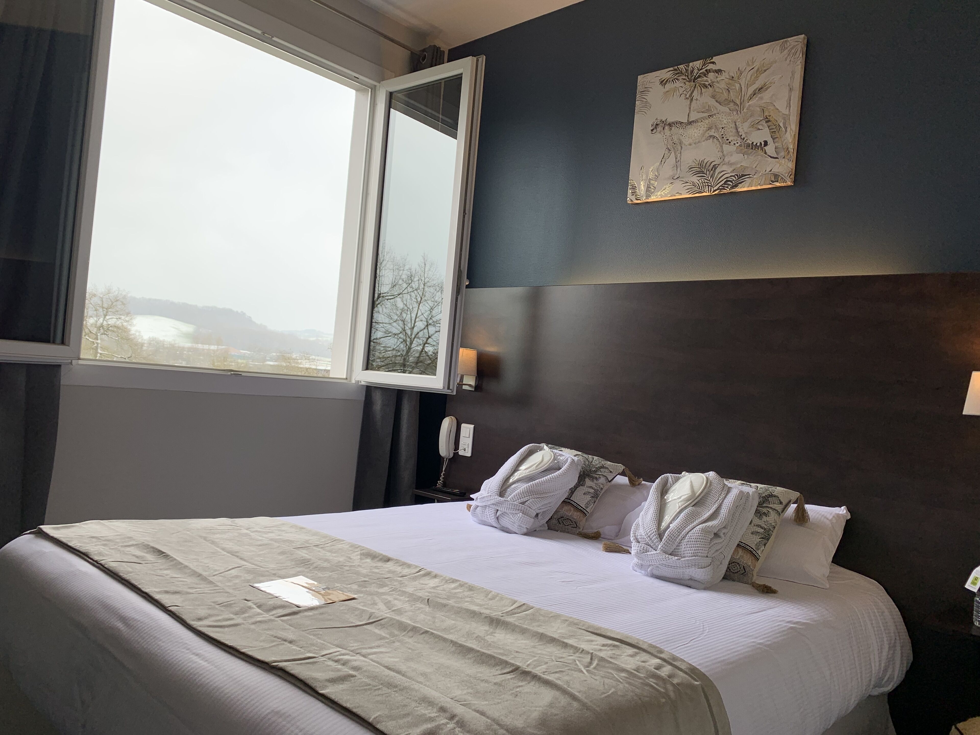 Une chambre d’hôtel avec un grand lit, une vue dégagée par une fenêtre ouverte, un tableau au mur et un téléphone sur la table de chevet.