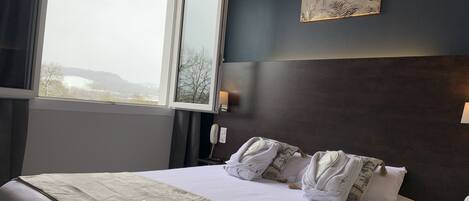 Une chambre d’hôtel avec un grand lit, une vue dégagée par une fenêtre ouverte, un tableau au mur et un téléphone sur la table de chevet.