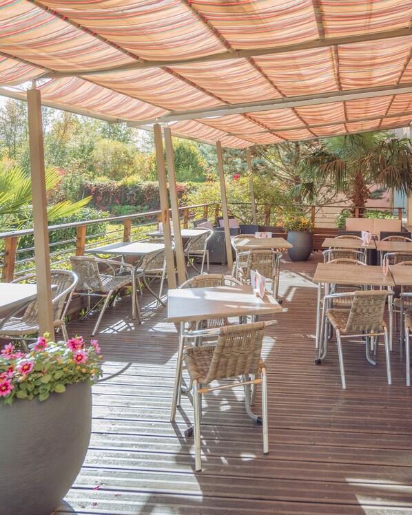 Une terrasse extérieure avec des tables et des chaises, entourée de verdure et surmontée d’un auvent à rayures.