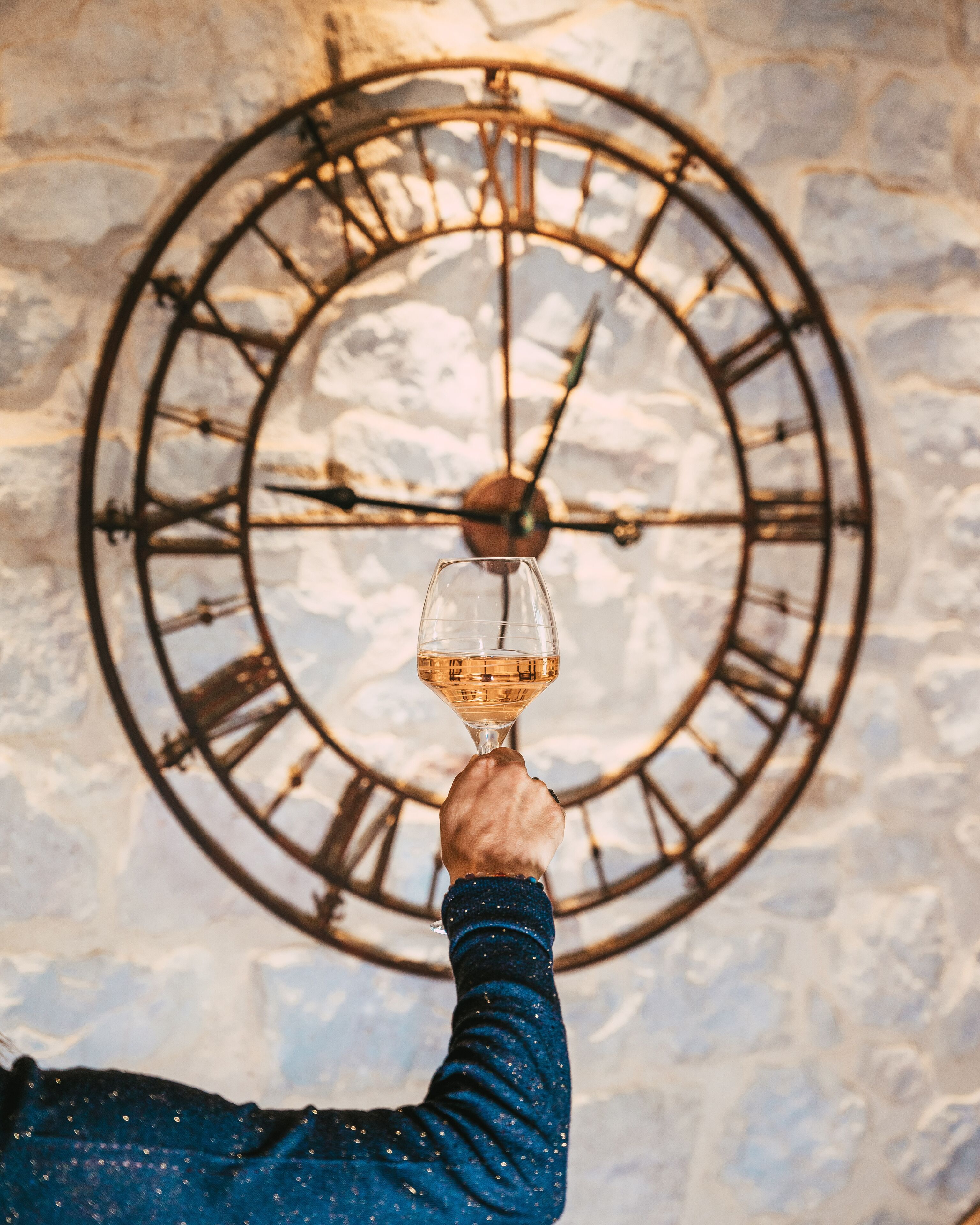 A hand holding a wine glass in front of a large, circular metal clock mounted on a stone wall.
