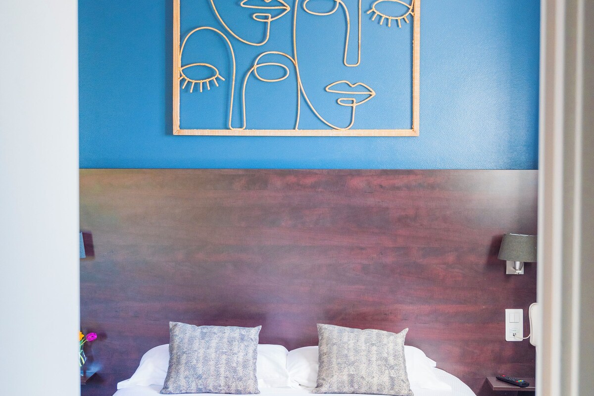 A hotel room with a bed, pillows, a wall art piece, a nightstand, and a lamp.