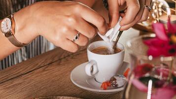Eine Person rührt ein Getränk in einer Tasse, die auf einem Untertasse steht. Auf dem Tisch befinden sich Gebäck und eine Blumenvase.