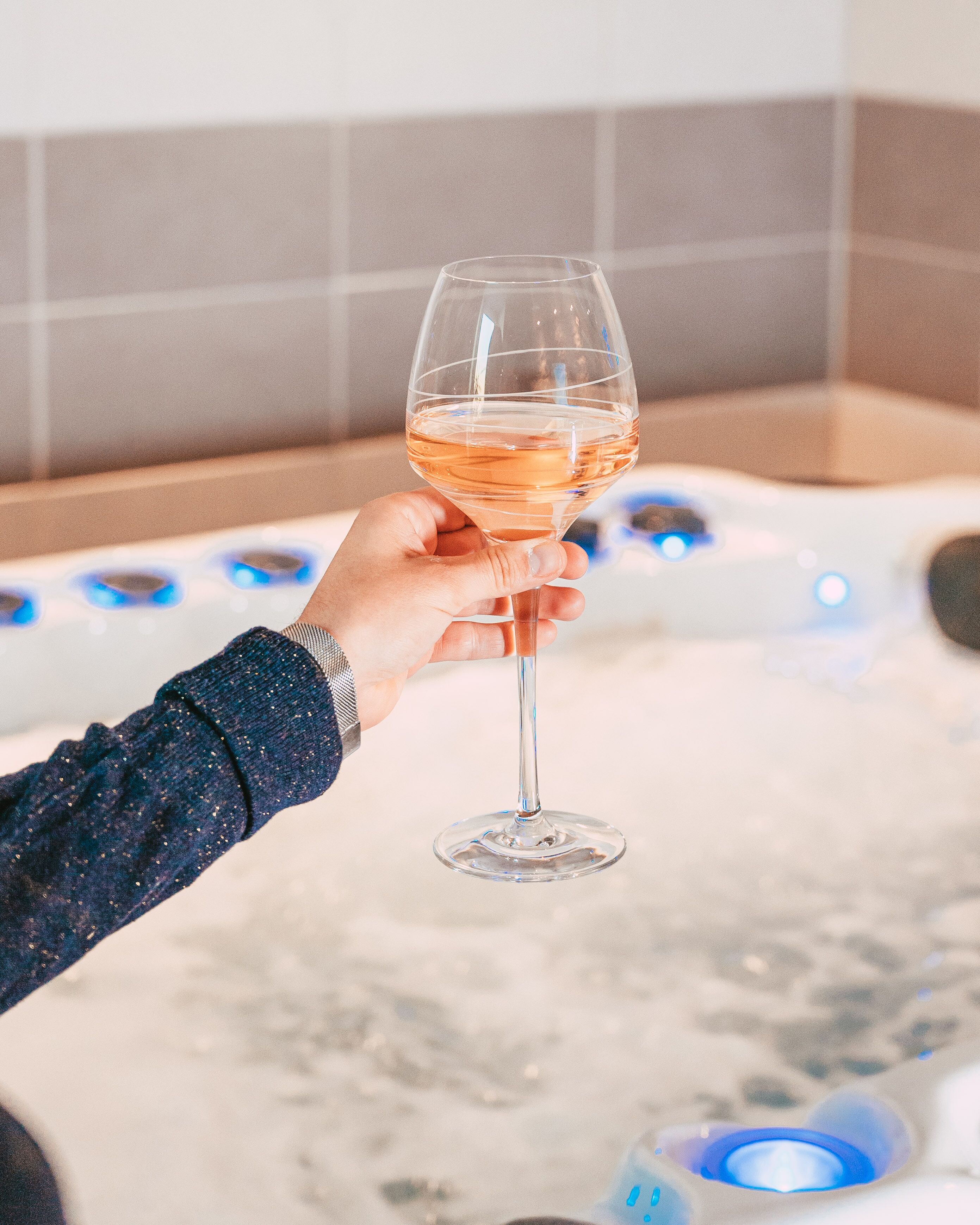 A hand holding a glass of rose wine in front of a hot tub with visible jets.