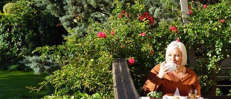A person sitting at an outdoor table with a cup and pastries, surrounded by lush greenery and flowering bushes.