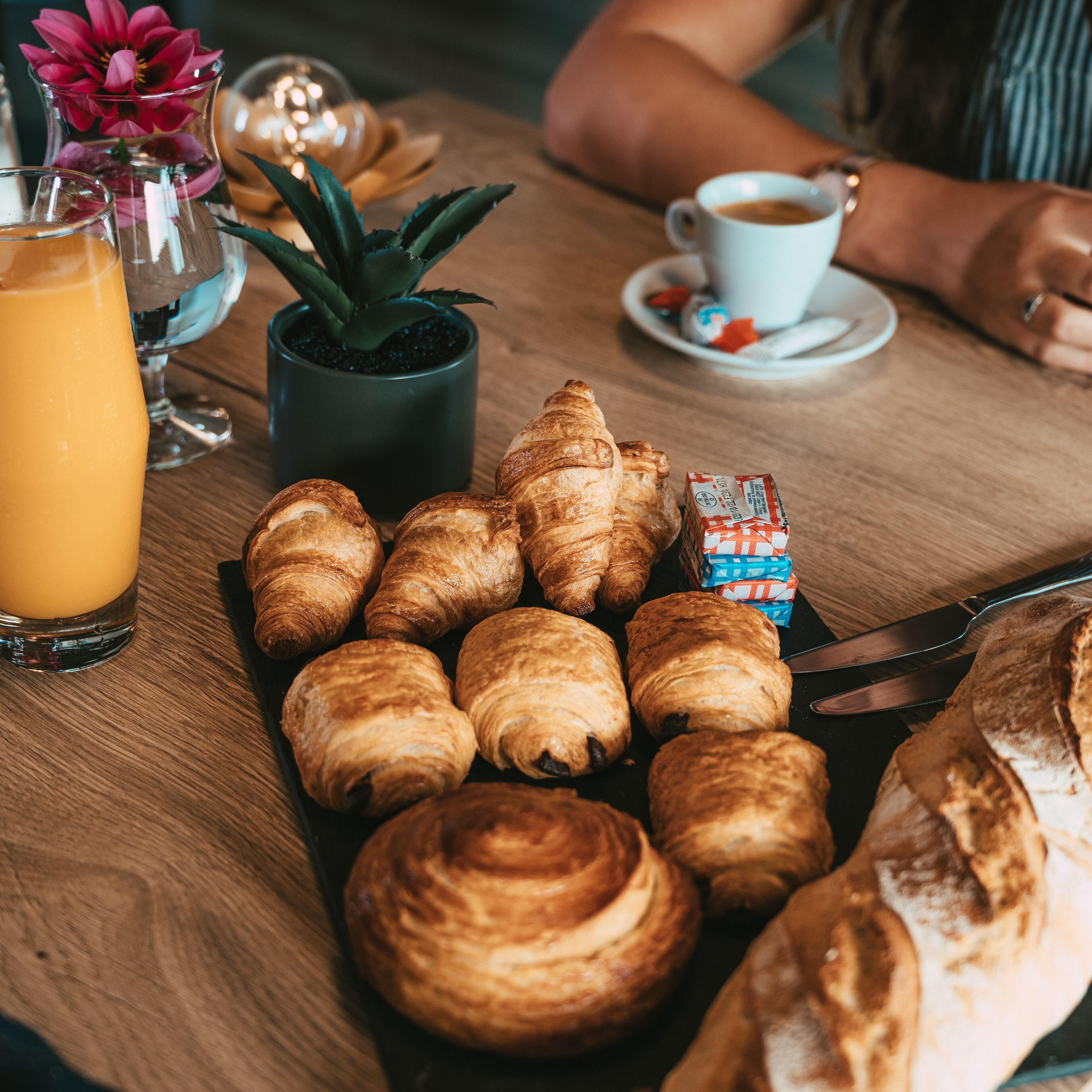 Une table en bois avec des croissants, un verre de jus d’orange, une tasse de café et une petite plante en pot.