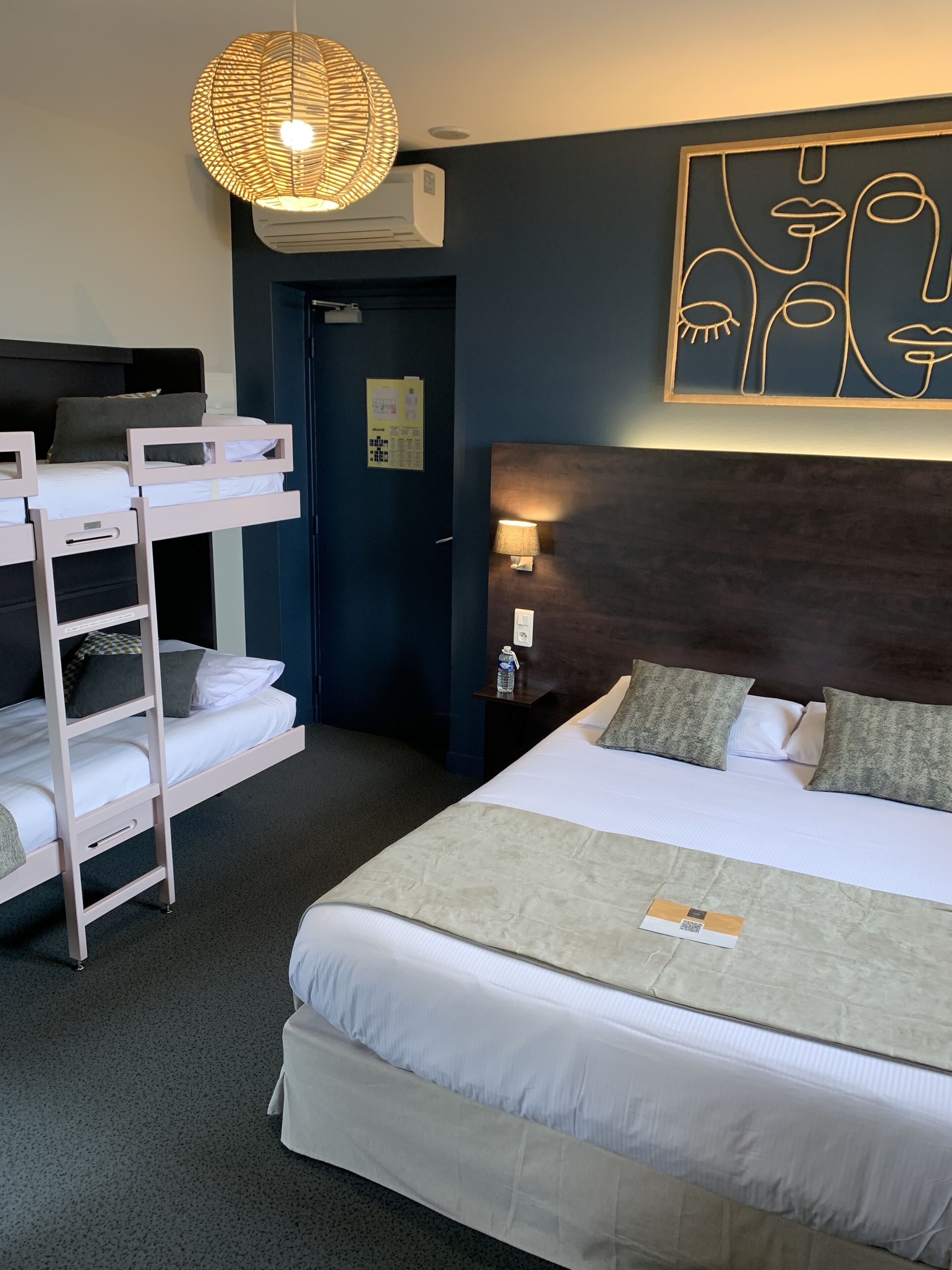 A hotel room with bunk beds, a large bed, a wall art piece, a lamp, and an air conditioning unit.