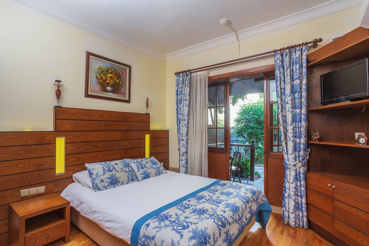 Un dormitorio con cabecera de madera, cama con colcha de flores azules y blancas, mesita de noche, televisor sobre una repisa de madera y una ventana con cortinas.