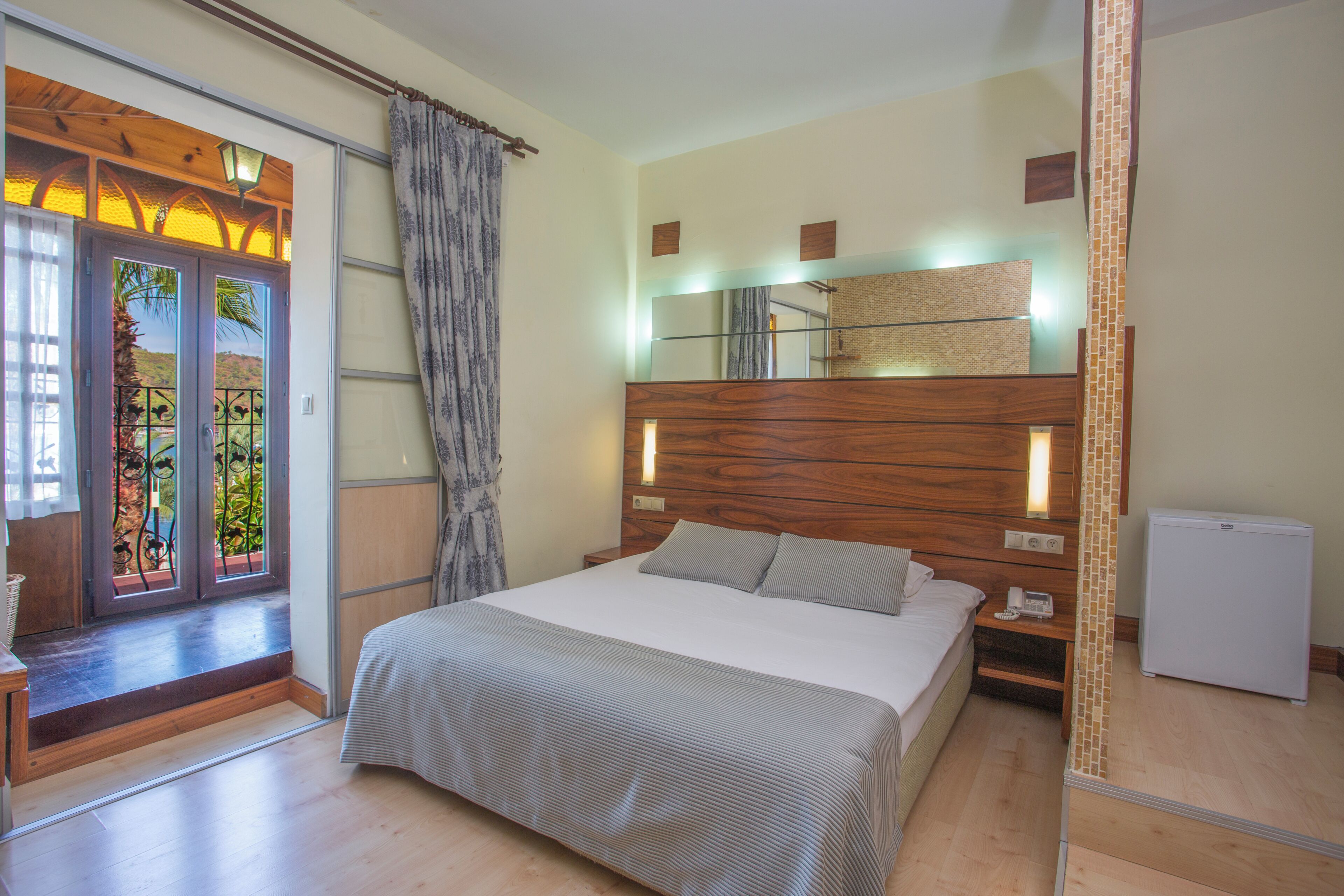 Une chambre à coucher comprenant un lit, des tables de chevet, une commode, un miroir, un petit réfrigérateur et une vue sur l’extérieur par une fenêtre.