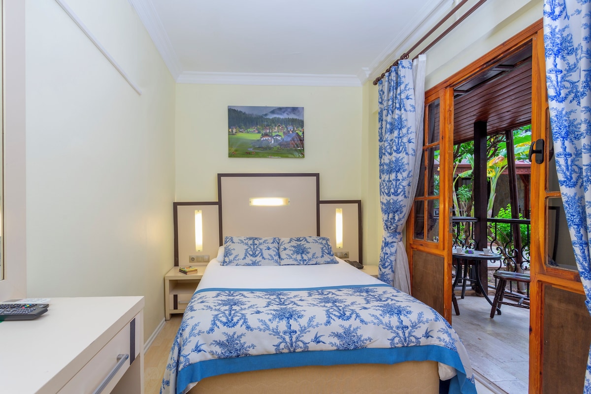 Un dormitorio con cama, mesitas de noche, un escritorio, un cuadro en la pared y vistas a través de un puerta abierta.