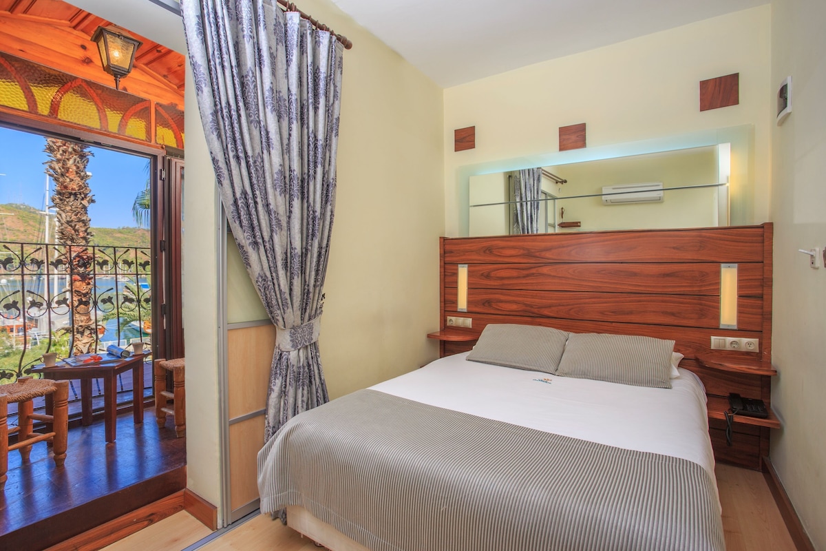 Habitación de hotel con una cama grande, cabecero de madera, una mesita y un balcón con vistas.