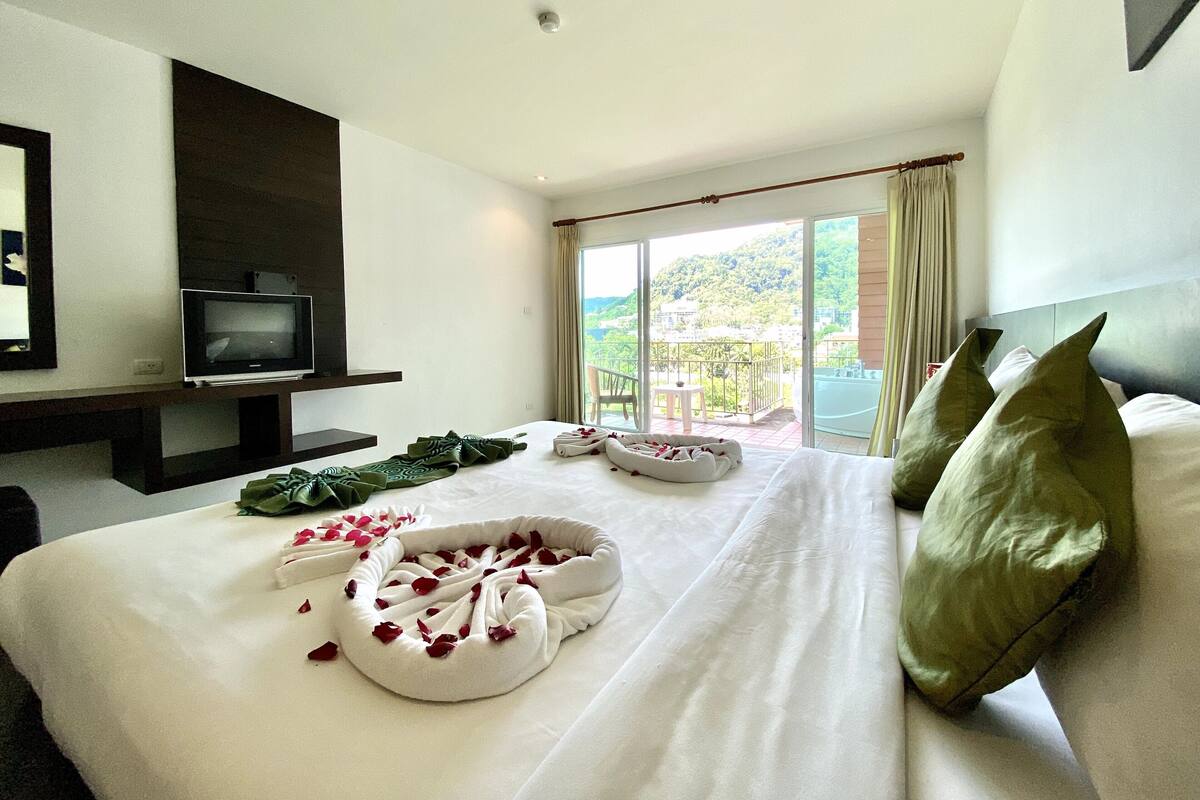 A hotel room with a bed, a TV, a balcony with a view of greenery, and a window with curtains.