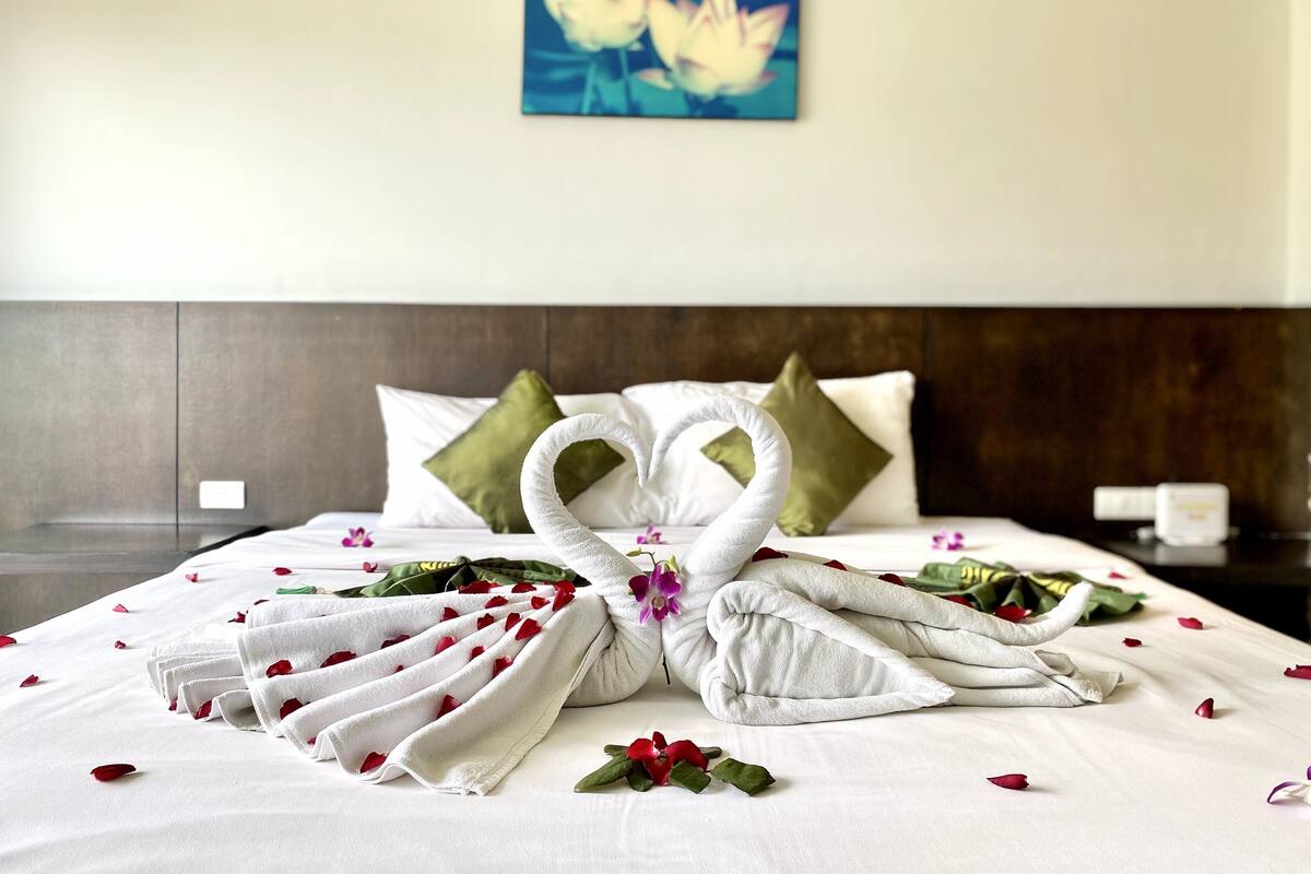 A neatly made bed with a towel sculpture, green pillows, and scattered rose petals.