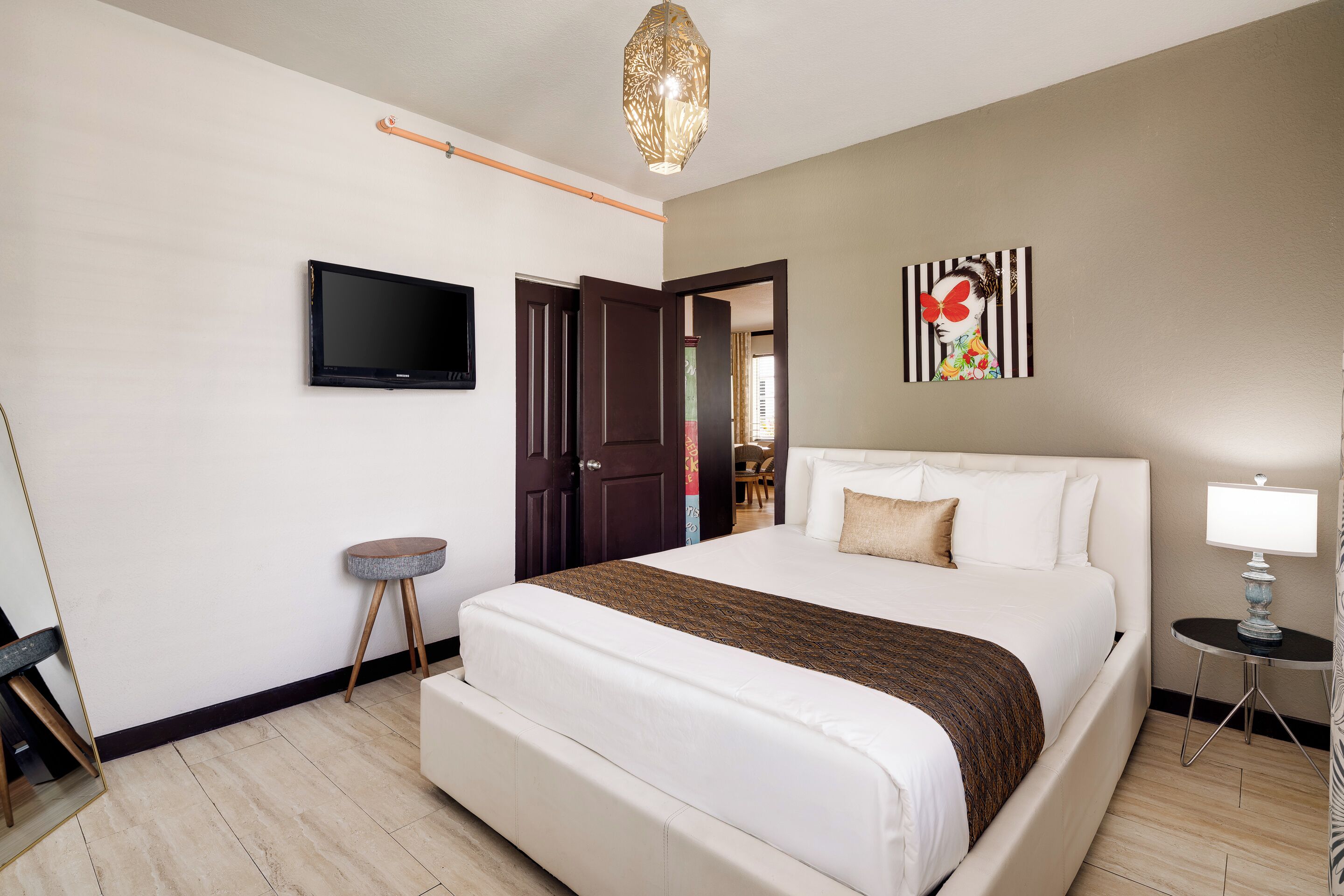 Habitación de hotel con una cama grande, un televisor de pantalla plana en la pared, una mesita redonda con una lámpara y una puerta que da a otra habitación.